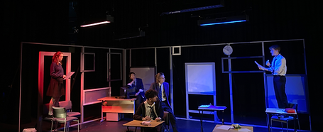 A wide photo of a classroom onstage, some students are stood on chairs. There is red and blue lighting. Photo by CaitlinBerry