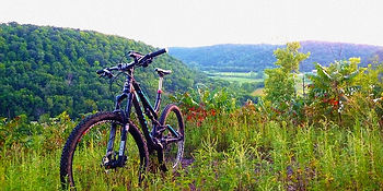 Mountain bike on a grassy hilltop overlooking lush green valleys and hills, under a light cloud cover.