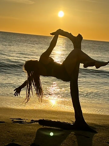 Tracy Jo standing on the beach, bathed in the warm, spiritual glow of a stunning sunset. Shades of deep orange, red, and pink fill the sky, blending seamlessly with the horizon, as the waves reflect the fiery colors and create a tranquil, almost otherworldly atmosphere.