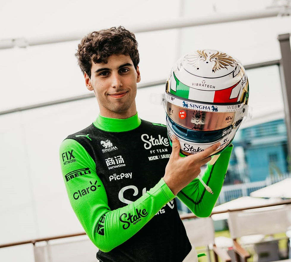 Piloto usa macacão verde e preto com patrocinadores. Segura na altura da cabeça um capacete com cores da bandeira da Itália