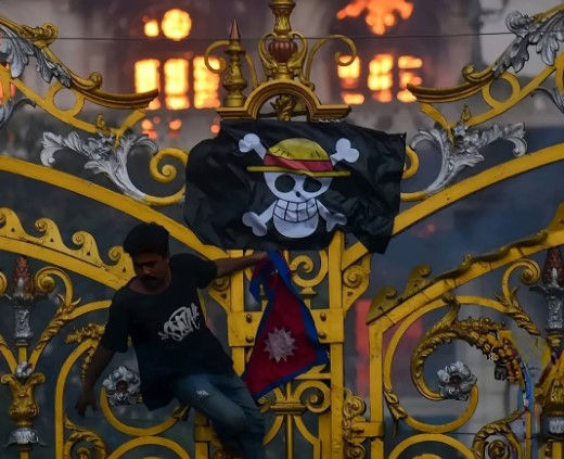 Homem pendurado em portão segurando bandeira do Nepal. Atrás está bandeira com símbolo pirata do Discord usado para escolher nova líder