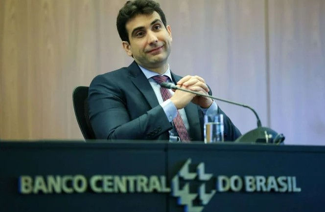 Gabriel Galípolo sentado atrás de mesa com nome e logo do Banco Central. Mãos cruzadas abaixo do queixo e terno de cor escura
