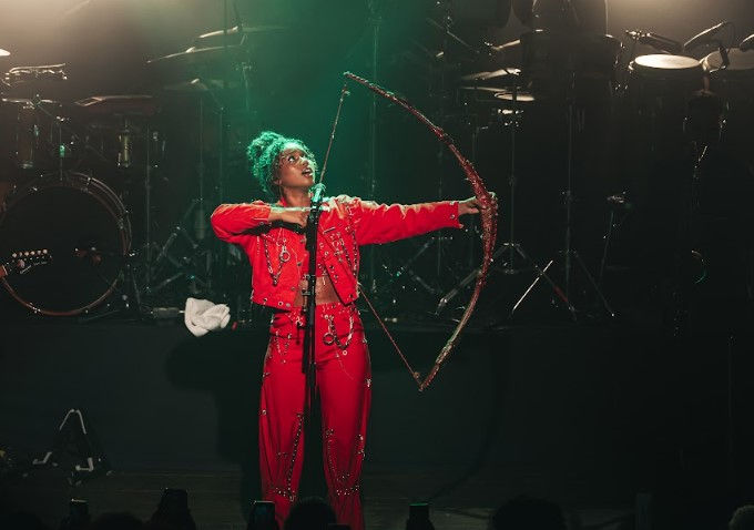 Cantora Melly vestindo vermelho, empunhando arco e flecha durante apresentação