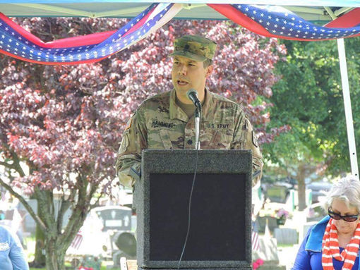 Soldier speaks at podium with text, cemetery in the background. Memorial service.