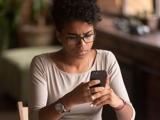 Woman with glasses looks intently at her smartphone indoors, wearing a beige top. Background is blurred, creating a focused, concerned mood.