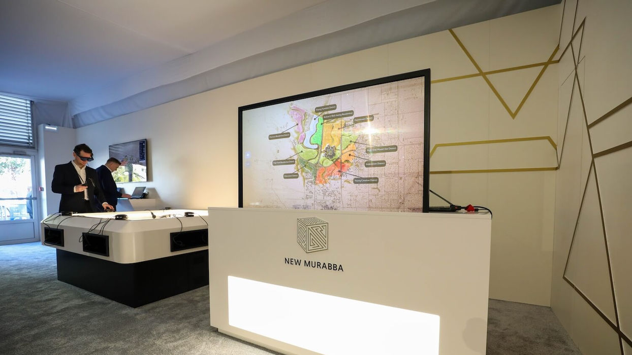 Two men interact with a large display screen reading NEW MURABBA, MIRRORLAB