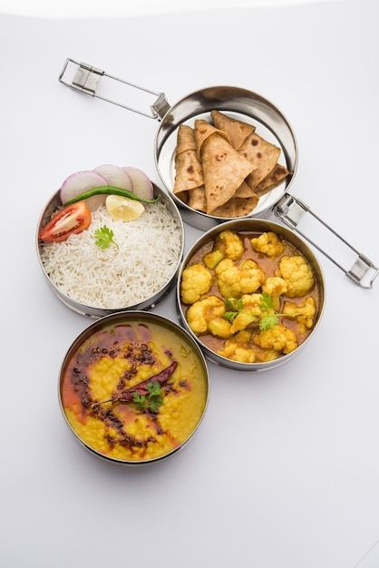 Metal lunch box with rice, bread, curry, and dal on white surface.