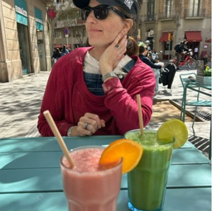 Picture of a woman with a baseball hat, sunglasses and a red sweatshirt sitting outside a cafe with a green and pink juice in front of her. 