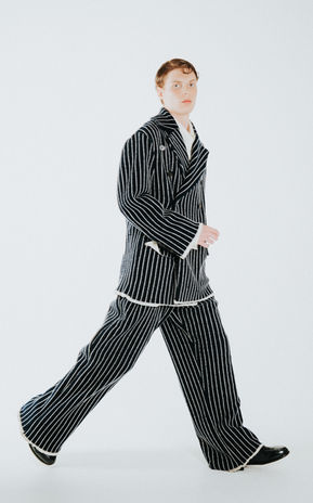 Editorial fashion image of a male model walking in a bold, oversized navy pinstripe suit with white trim. The makeup look is fresh and clean with a luminous base, soft contouring, and a natural flush—ideal for high fashion menswear or runway grooming campaigns.