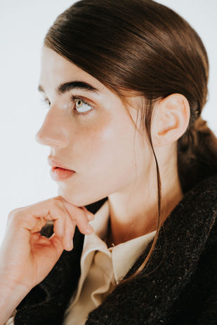 Close-up profile of a model showcasing minimalist fashion makeup—dewy skin, natural flushed cheeks, brushed-up brows, soft nude lips, and subtle highlight. Hair is sleek and parted with a modern wet-look finish, ideal for editorial or runway styling
