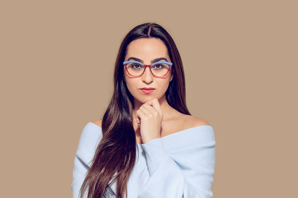 Woman with long hair and colorful glasses, wearing a light blue off-shoulder sweater, poses against a beige background, appearing thoughtful.
