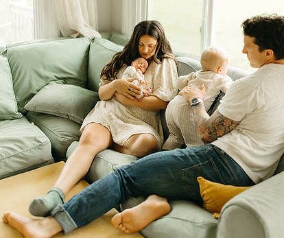 Founder Julianna sits in her sunroom with husband and two children - a toddler looks out the window and a newborn is being shushed to sleep