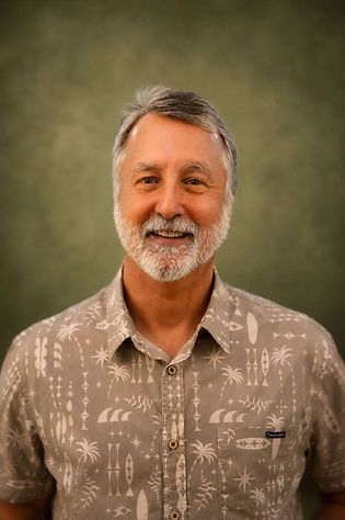 Smiling man with gray short hair and a white beard wearing a brown and tan print button up shirt.