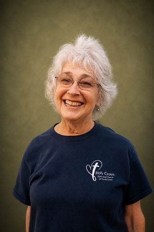 Brightly smiling woman with short, fluffy white hair and glasses with clear frames wearing a black t-shirt with Holy Cross design.