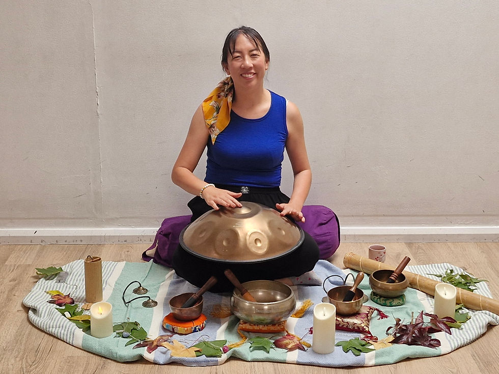 Smiling woman sits on a blanket playing a bronze handpan, surrounded by candles, singing bowls, leaves, and other meditation items.