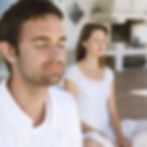 Man and woman in white sit with eyes closed on a sunny porch, शांत and meditating in a calm, airy setting