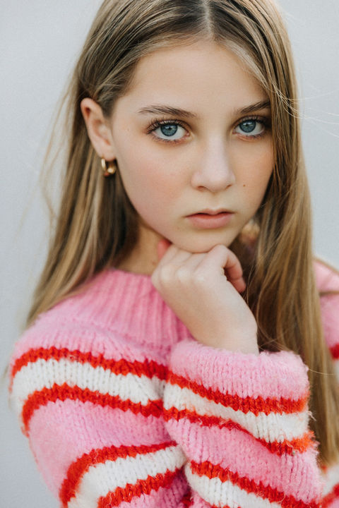 Headshot of teen model with long dark blonde hair wearing a pink jumper with red and white stripes. Resting her chin on fisted hand.