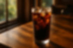Glass of iced coffee with ice cubes on a wooden table, warm sunlight streaming through a window, creating a cozy ambiance.