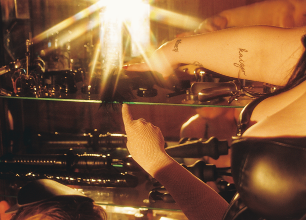 Close-up of a Dominatrix's tattooed arm reaching across a glass shelf of fetish toys to show Her sissy maid where to clean