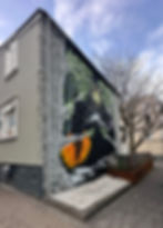 Street art mural on a gray building, featuring colorful trees and a moon. A bare tree stands beside the building under a cloudy sky.