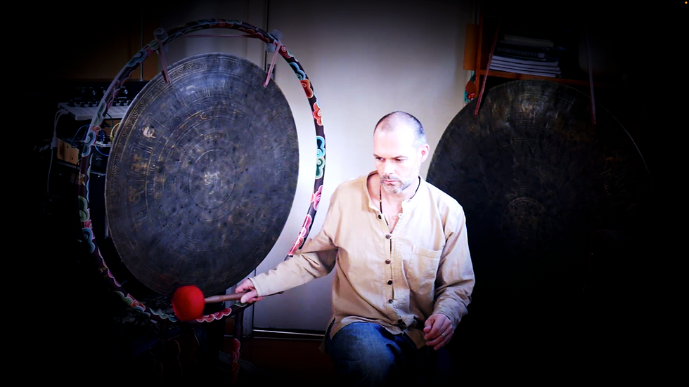 A person plays a large gong with a red mallet in a dim room. Two large gongs with colorful frames are in the background, creating a serene mood.
