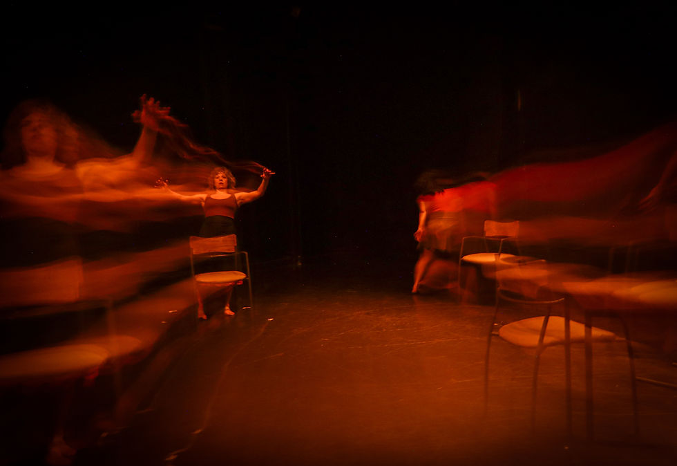 Bailarines borrosos en un escenario oscuro con sillas, iluminados en rojo y naranja, creando un ambiente dramático. Atisbos de un ocaso