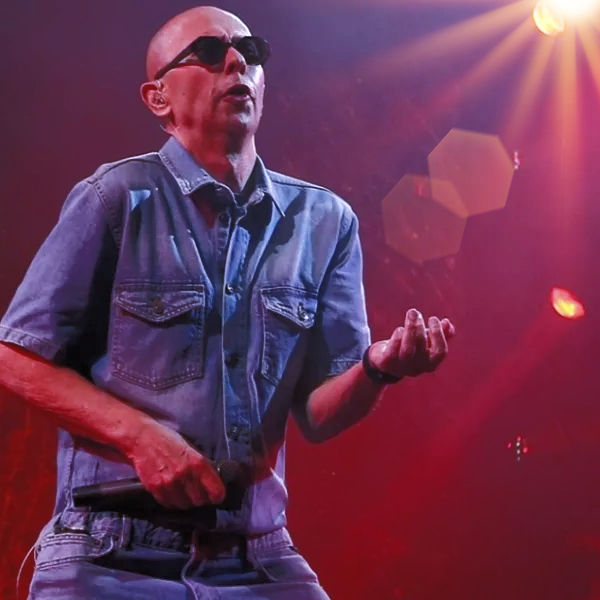Cantante calvo con gafas de sol y camisa vaquera, con micrófono en mano, actuando en un escenario rojo y morado. El indio Solari