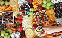 A vibrant snack charcuterie board featuring crackers, cheese, nuts, fresh fruit, chocolate, and candy, with a child’s hand reaching for a sweet treat.