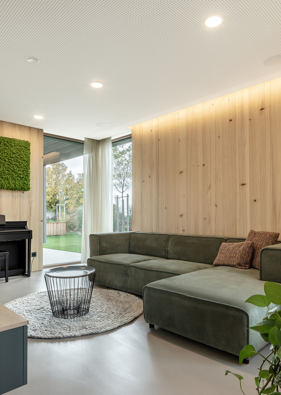 Gemütliches Wohnzimmer mit hellen Holzwänden, einem großen grünen Ecksofa, indirekter Deckenbeleuchtung und bodentiefen Fenstern.