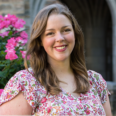 Rev. Kelsey Cobbs smiles at the camera wearing a floral printed dress and wavy light brown hair with pink flowers in the background.