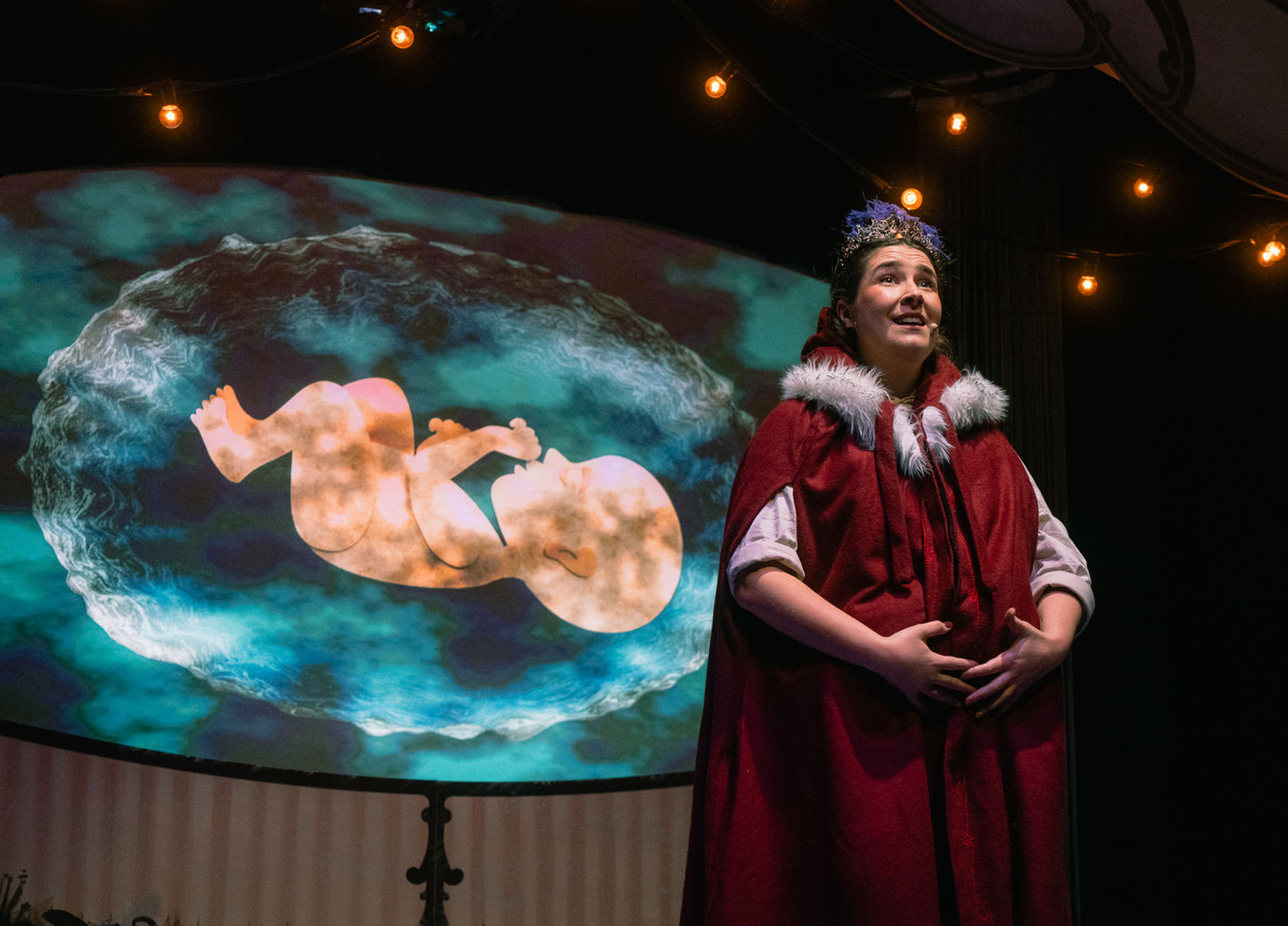 Smiling actress in red cloak, fetus projection behind her