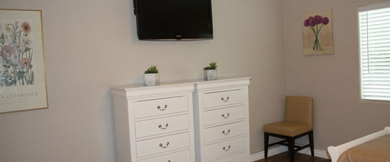 Bedroom with two white dressers, wall-mounted TV, and small chair.