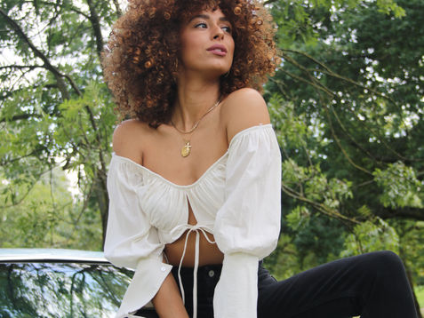 A woman with curly hair in a white off-shoulder top and black trousers sits on a light blue vintage car, posing confidently under a canopy of trees.