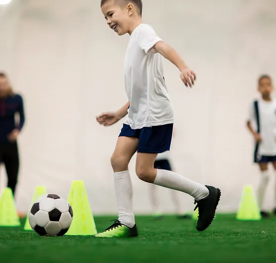 Happy boy in uniform kicking the ball while running around cones during training.jpg.webp