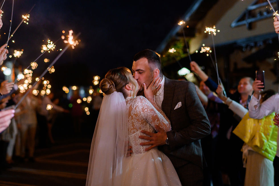 Bride and groom kiss in a sparkler send-off at night, surrounded by guests holding lit sparklers and cheering at a wedding.
