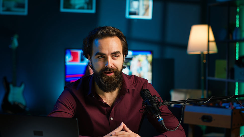 Bearded man in a maroon shirt speaks into a podcast microphone in a dim blue-lit studio with screens and a lamp.