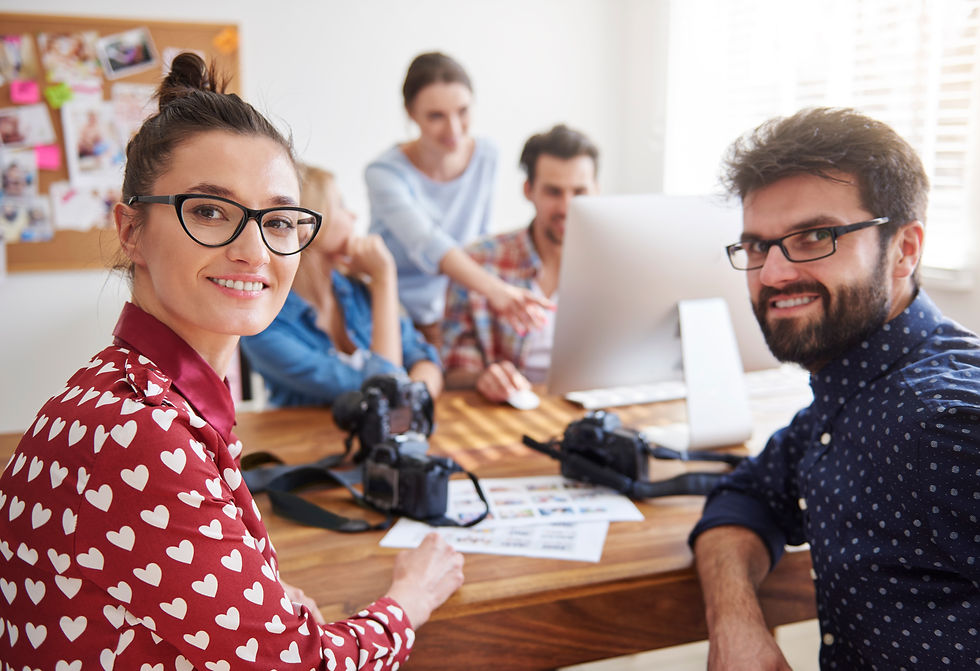 Smiling coworkers in a bright office gather around a desk with cameras and a computer, looking at the viewer.