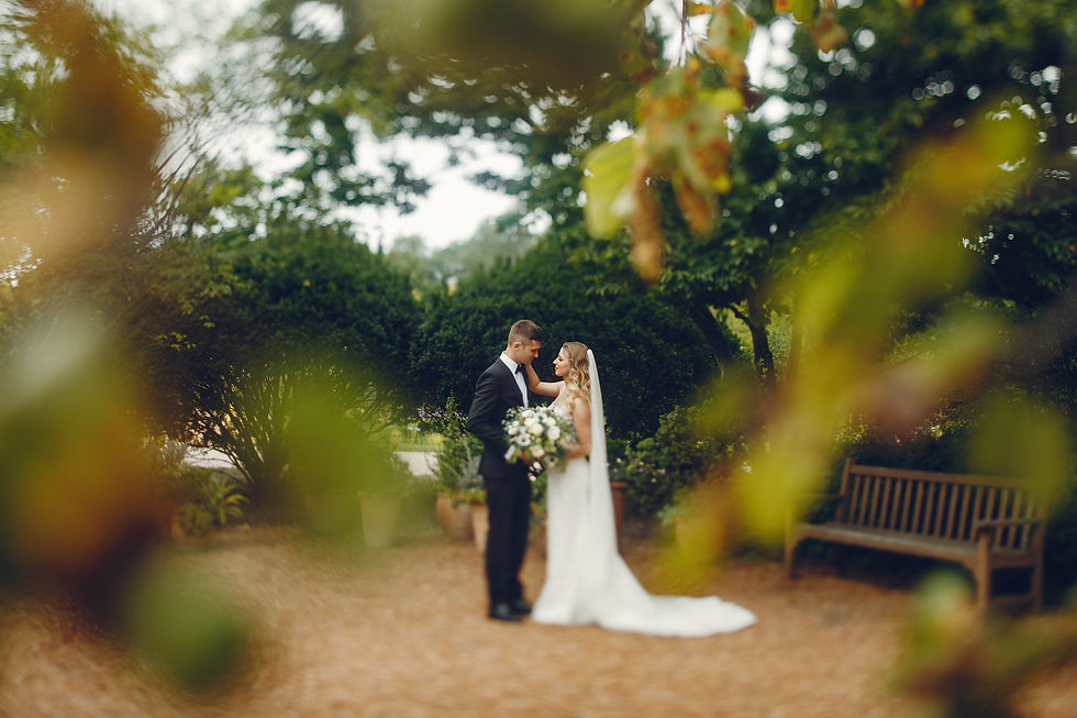 Wedding couple embrace in a lush garden, bride in white gown holding bouquet, groom in black suit, with blurred leaves foreground.