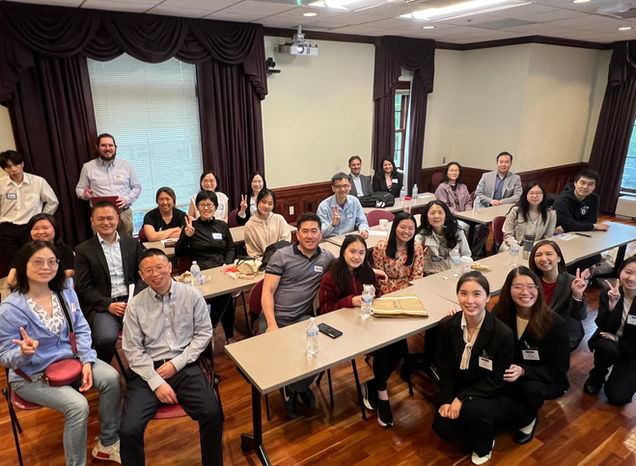 A group photo taken in a classroom or conference room setting featuring around 30 people, mostly seated at tables. The attendees appear to be a mix of students and professionals, with many smiling and showing peace signs. Some have name tags, and a few carry bottled water or notepads. The atmosphere seems friendly and relaxed, possibly from a seminar, workshop, or networking event. The room has wood flooring, maroon drapes, and a ceiling-mounted projector.
