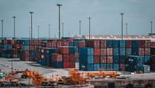 Stacks of blue and red shipping containers at a port, with cranes and light posts under a cloudy sky. Some containers bear visible text.