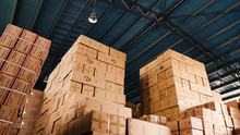 Tall stacks of taped cardboard boxes in a warehouse under a blue metal roof, with labels like WHITE and BLACK.