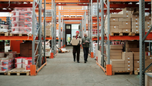 Two warehouse workers carry a cardboard box down a shelving aisle lined with stacked boxes and paint buckets.