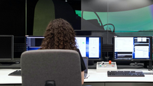 Person with curly hair at control center desk, facing multiple monitors displaying data in a dimly lit room.