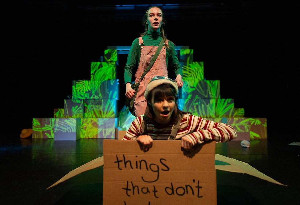 A performer pops up from inside a large cardboard box, holding a hand-drawn sign that begins “things that don’t...”. Another performer stands behind, looking out, in front of a leafy green-projected backdrop.
