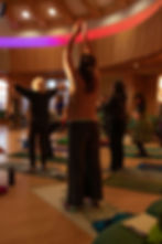 People engage in yoga in a warmly lit room at the Medicine Womyn retreat in Vermont, arms raised, on patterned mats. A colorful ceiling adds a serene ambiance.
