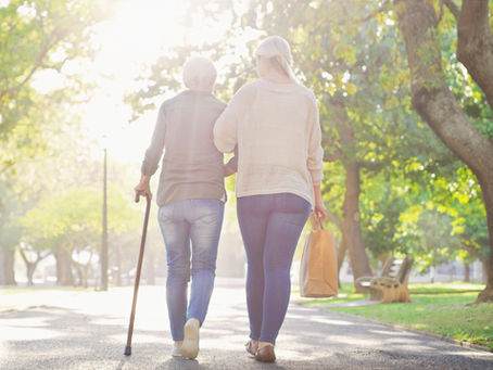 A senior mother, using a cane, strolls arm in arm with her adult daughter, their backs to us, on a sunny autumn day.