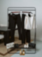 Capsule wardrobe hanging on a black clothing rack featuring a white tank top, light blue button-down shirt, plaid blazer, dark trousers, and black Prada handbag with heels.