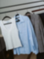 Curated capsule wardrobe essentials hanging on a minimalist rack, featuring a white ribbed top, light blue button-up, and tailored blazer. Image links to Services Page.