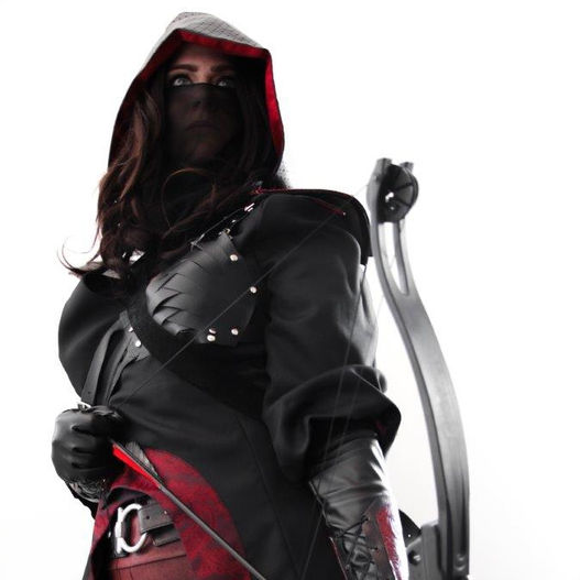 A woman in a black coat with burgundy trim and leggings and a black hood and veil wields a bow and arrow