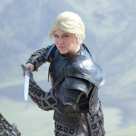 A woman with short blonde hair wields a sword, wearing full plate armor and a chainmaille-covered tunic